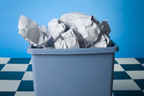 A close-up image of a light grey plastic bin filled with crumpled white paper waste, situated on a black-and-white checkered floor with a solid bright blue background. The crumpled paper varies in size and shape, with some pieces spilling over the top of the bin, which is positioned slightly off-center. The setting appears to be an indoor environment suitable for rubbish collection or disposal. The visual focus is on the paper waste in the bin, highlighting the textures of the crumpled paper and the smooth surface of the plastic container, emphasizing the importance of proper rubbish removal services in local areas such as Gerrards Cross. The image, captured with clear lighting, underscores the type of waste managed by companies like House Clearance Gerrards Cross, connecting the visual content subtly to rubbish removal efforts around the region.