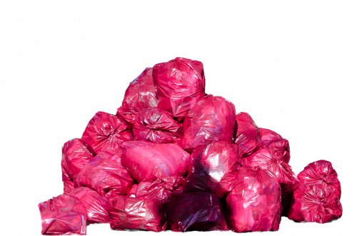 A large pile of pink plastic rubbish bags, tightly sealed and stacked outdoors on a flat surface. The bags vary slightly in size and shape, with some showing creases and wrinkles in the plastic material. The background is plain white, with no other objects or environmental features visible. The bags appear to contain general waste or rubbish, commonly associated with household or garden rubbish removal services. The image suggests a collection ready for disposal or rubbish clearance work, typical of services provided by companies like House Clearance Gerrards Cross. The pink colour of the bags adds a vibrant contrast to the overall scene, which is neutral and professional in tone. The orderly stacking of the bags indicates they are prepared for transport, fitting the context of rubbish removal in a residential area in or near Gerrards Cross.