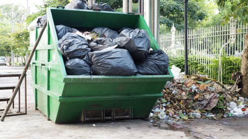 A green industrial skip container filled with tightly packed black rubbish bags made of plastic, located outdoors on a paved surface, with some loose waste and discarded paper or packaging materials scattered beside it. The skip is positioned adjacent to a nearby fence and surrounded by trees and shrubs, indicating a residential or park area in Gerrards Cross. The environment appears to be a typical rubbish collection site, with natural light illuminating the scene, emphasizing the utilitarian nature of rubbish removal services provided by House Clearance Gerrards Cross. The image captures the process of waste temporarily stored for collection, highlighting the outdoor setting, the contrast between the green container and black bags, and the cluttered surroundings, which are consistent with local rubbish disposal or clearance activities in the area.
