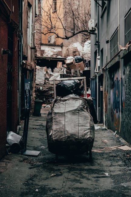 A large collection of mixed household rubbish and discarded packaging is piled on a paved area in front of wheelie bins, including a gray recycling bin overflowing with crumpled paper, cardboard boxes