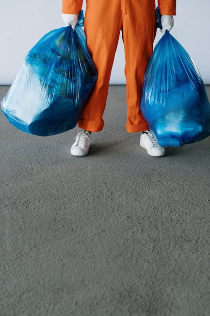 Close-up view of an individual wearing an orange work suit and a white glove, holding a large blue plastic rubbish bag with a crinkled texture and semi-translucent appearance. The person is gripping t
